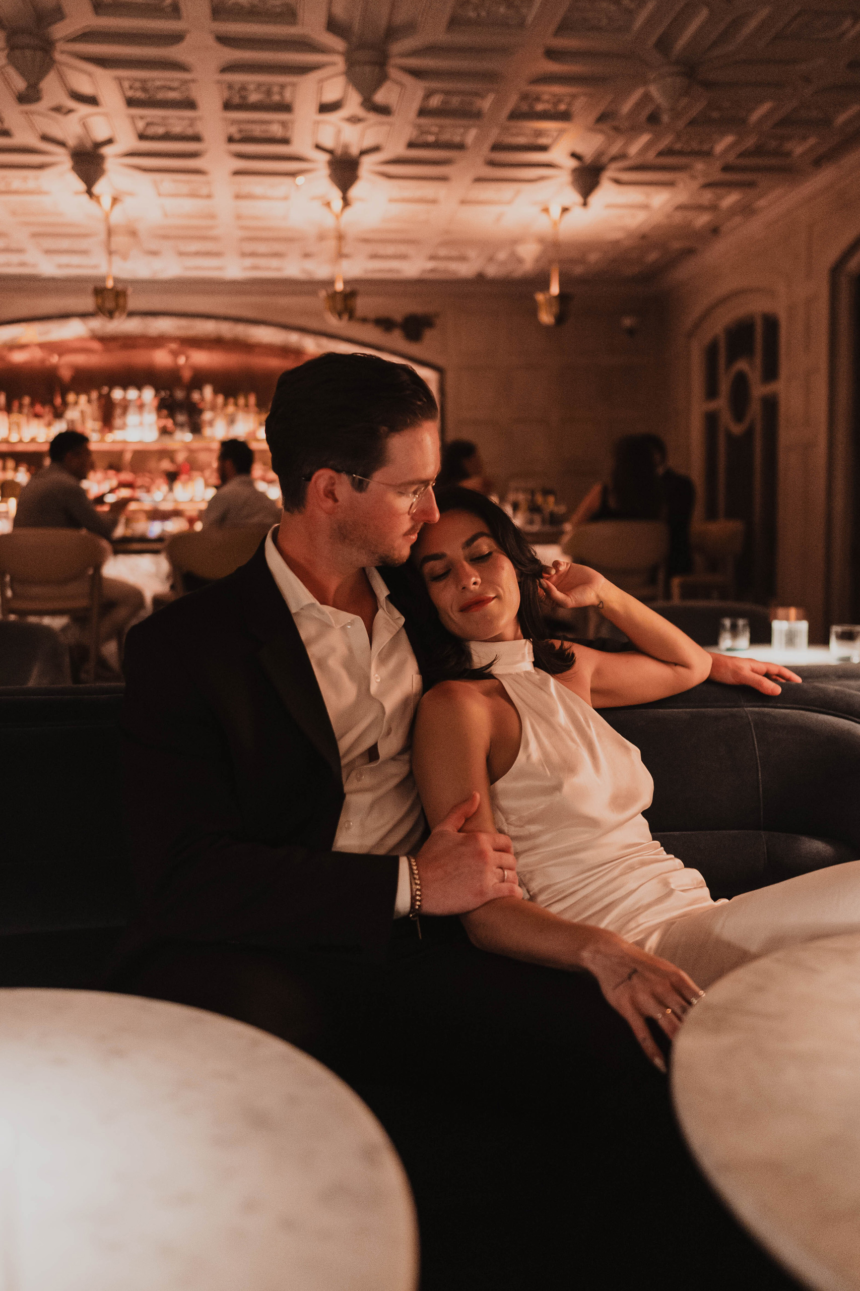 A stylish couple relaxing together on a velvet booth in a warm, candlelit lounge/bar setting.
