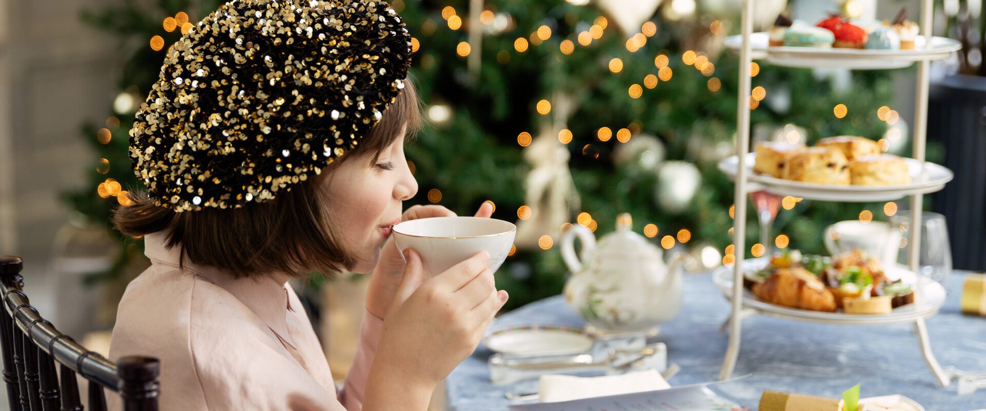 a young girl in holiday dress sipping tea near a Christmas tree