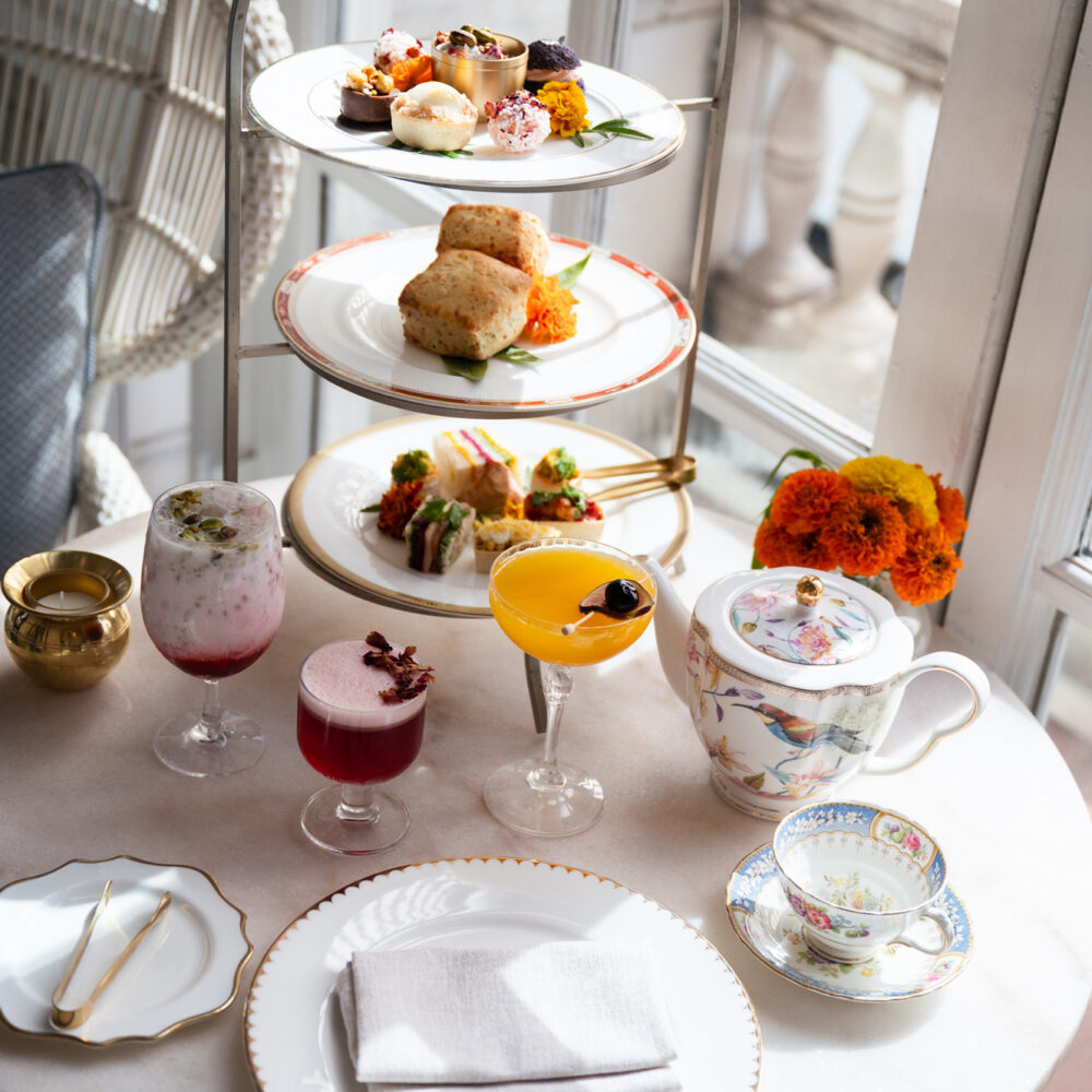 Afternoon tea spread with tiered pastries, colorful drinks, a floral teapot, and marigolds.