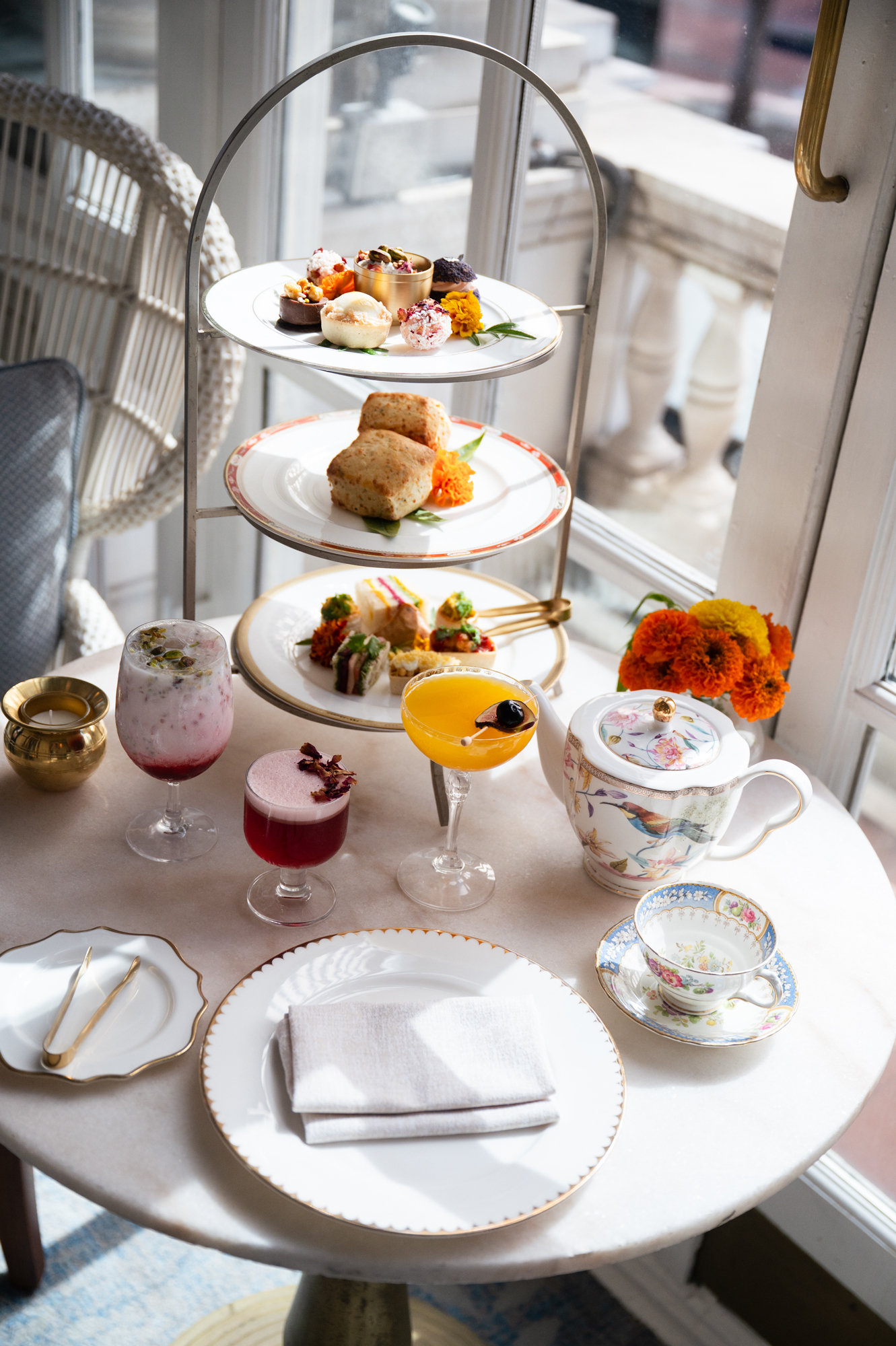 Afternoon tea spread with tiered pastries, colorful drinks, a floral teapot, and marigolds.