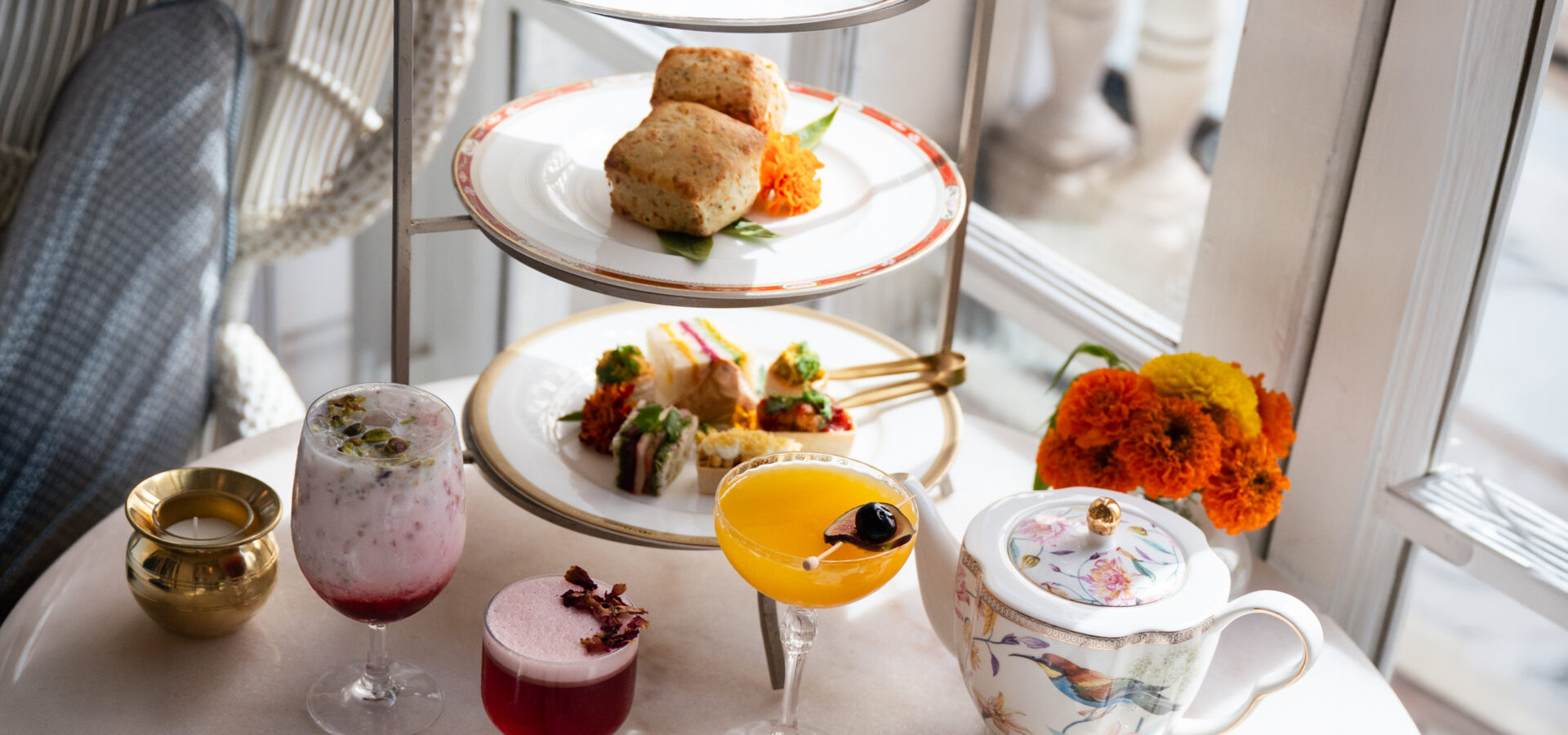 Afternoon tea spread with tiered pastries, colorful drinks, a floral teapot, and marigolds.